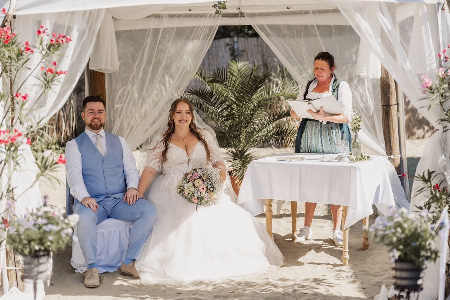Jessica und Jonas sitzen lachend unter dem weißen Baldachin bei ihrer freien Trauung am Sandstrand im Antik in Nittenau