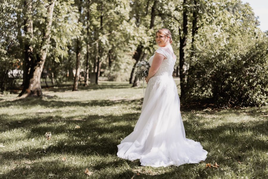 Christina in ihrem Brautkleid unter Bäumen im Park, sie schaut lächelnd über die Schulter