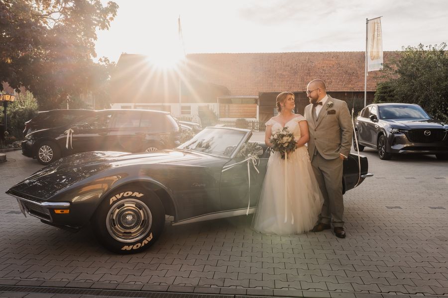 Christina und Dominik stehen im Gegenlicht der Abendsonne neben Christinas geschmückter dunkelgrüner Corvette vor dem Gasthof Koller