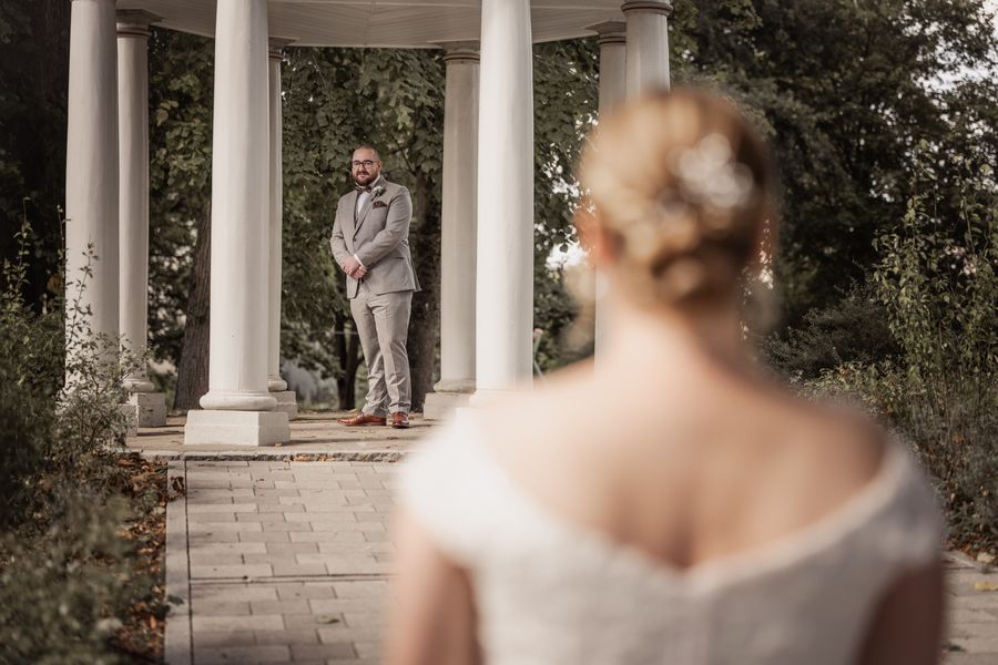 First Look: Dominik wartet im weißen Säulenpavillon, im Vordergrund unscharf Christina von hinten